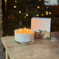 A lit scented candle in a clear glass jar sits on a rustic wooden table next to its product box. The background is a warm, softly blurred indoor setting with a glowing Christmas tree and warm lights.