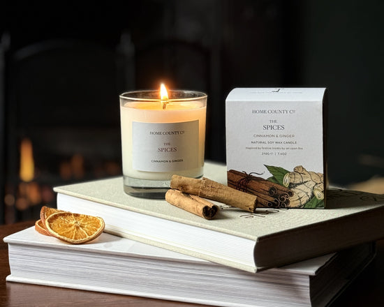 The cinnamon and ginger scented Christmas candle from the Home County Co. is shown alight next to its candle box with cinnamon sticks and orange slices in the foreground and an open fire in the background