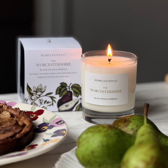 The Worcestershire Black Pear and Freesia scented candle from Home County Co is shown alight next to its candle box with fresh pears in the foreground