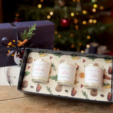 A three-scented scented candle gift set from Home on the Hill, displayed in an open decorative box with a matching purple gift box behind it on a wooden table, with a blurred Christmas tree in the background.