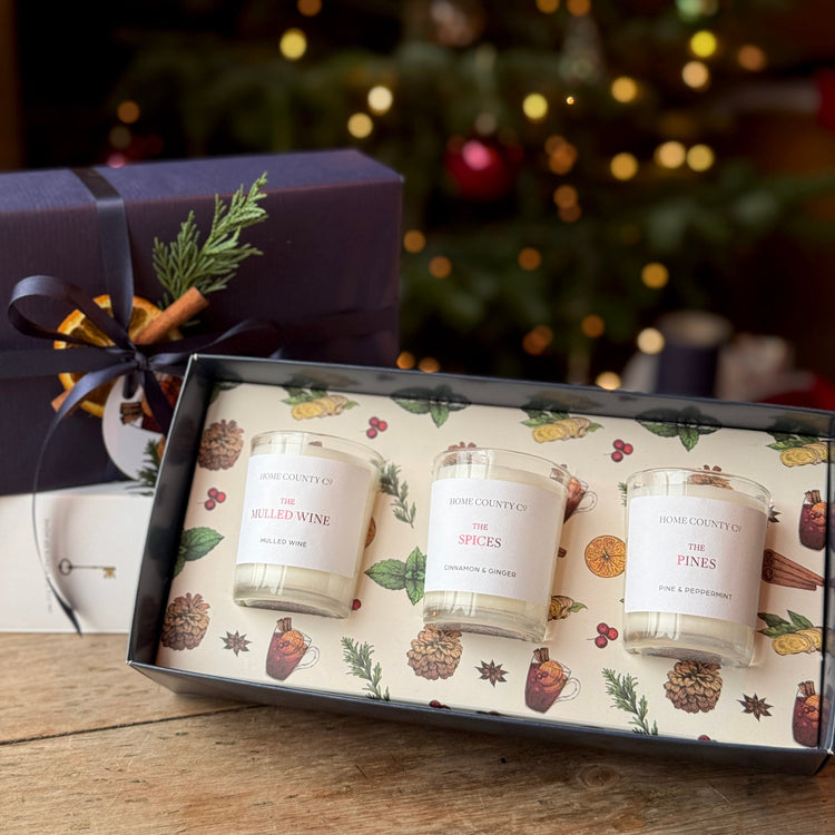 A three-scented scented candle gift set from Home on the Hill, displayed in an open decorative box with a matching purple gift box behind it on a wooden table, with a blurred Christmas tree in the background.