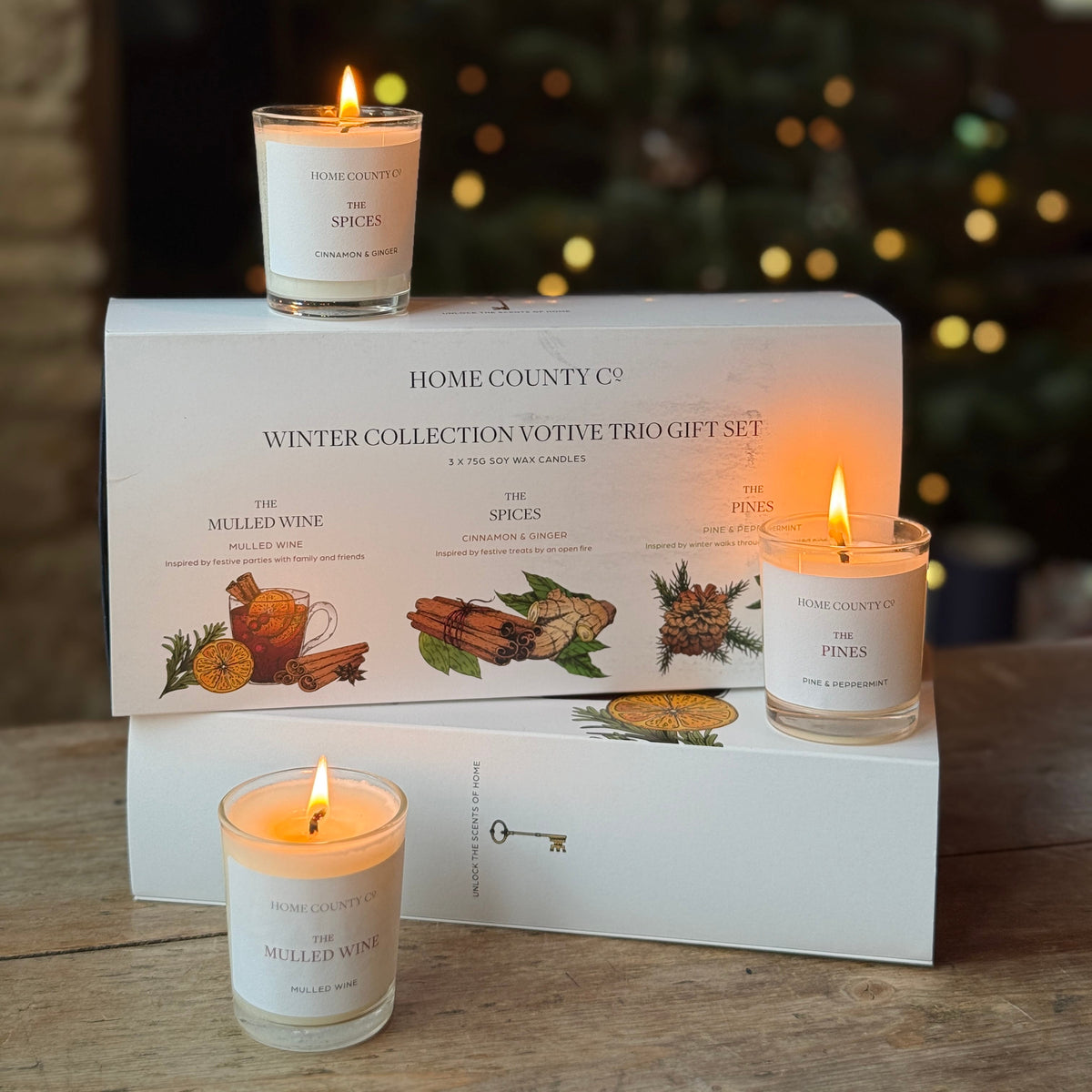 Home County Co. Winter Collection Votive Trio Gift Set featuring three lit white candles in glass jars arranged on top of and in front of white gift boxes on a wooden table, with a blurred Christmas tree in the background.