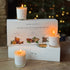 Home County Co. Winter Collection Votive Trio Gift Set featuring three lit white candles in glass jars arranged on top of and in front of white gift boxes on a wooden table, with a blurred Christmas tree in the background.
