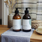 A 300ml coastal sea salt and wood sage scented hand wash and lotion duo from the Home County Co. is shown in eco-friendly amber glass bottles in a bathroom
