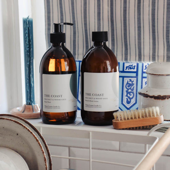 A coastal sea salt and wood sage scented liquid hand wash and hand and body lotion refill are shown on a kitchen windowsill in their eco-friendly amber glass bottles