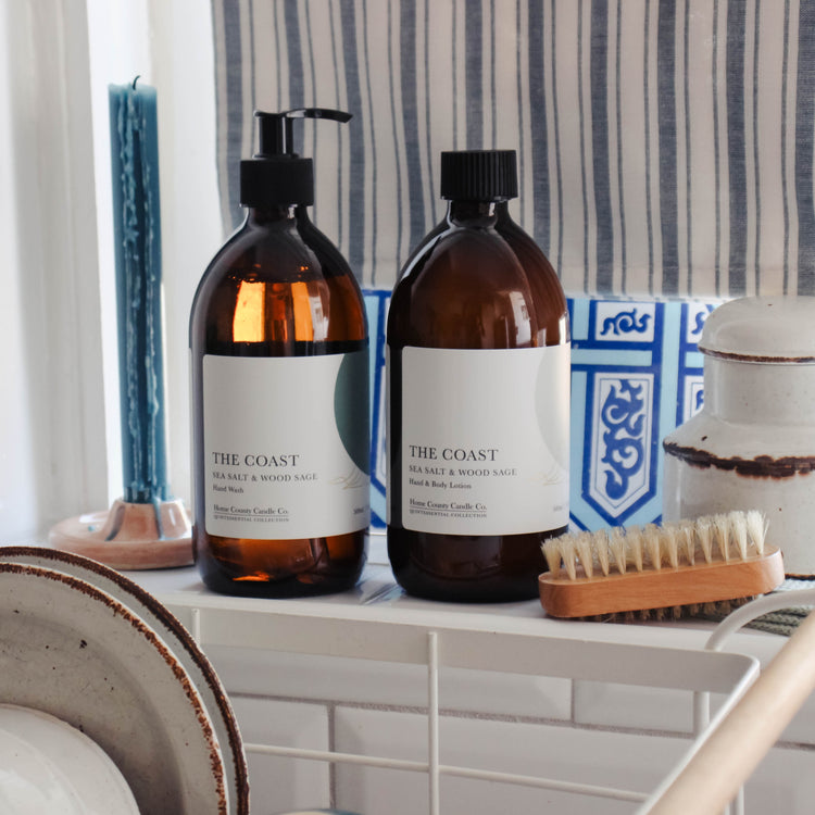 A coastal sea salt and wood sage scented liquid hand wash and hand and body lotion refill are shown on a kitchen windowsill in their eco-friendly amber glass bottles