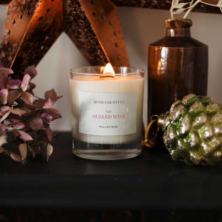 A Mulled Wine scented candle from Home County Co. is shown lit on a mantelpiece of Christmas decorations