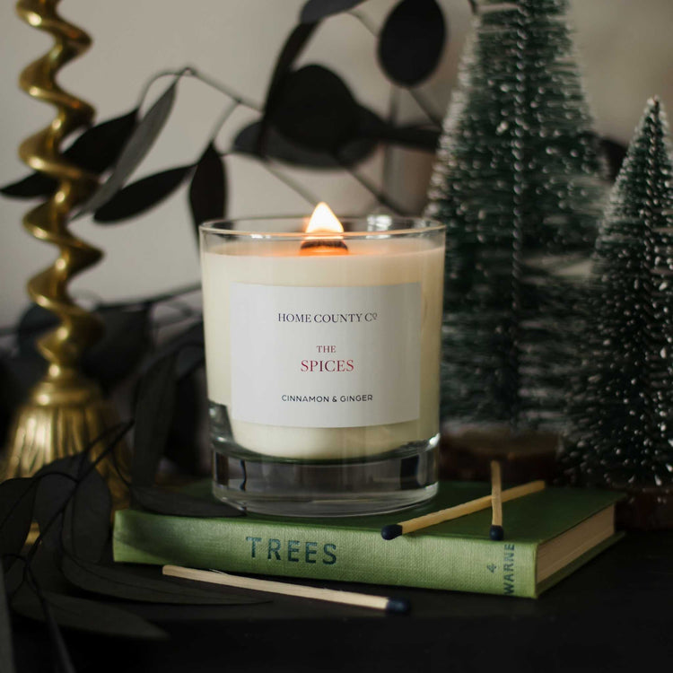 A cinnamon and ginger scented christmas candle from the Home County Co is shown with christmas tree decorations and the wooden wick lit.