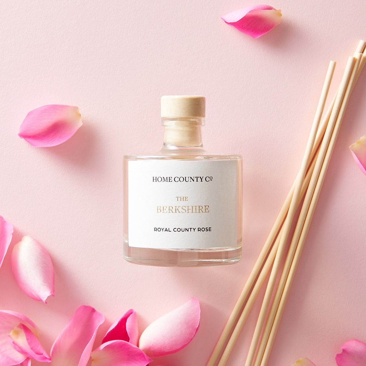A Berkshire county rose scented reed diffuser from the Home County Co. is shown next to fresh pink rose petals and porex reeds