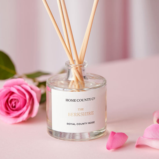 A Berkshire county rose scented reed diffuser from the Home County Co. is shown next to a fresh pink rose