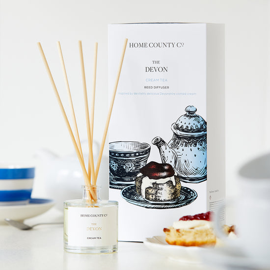 A Devon cream tea scented reed diffuser from the Home County Co. is shown next to its eco-friendly illustrated reed diffuser  box