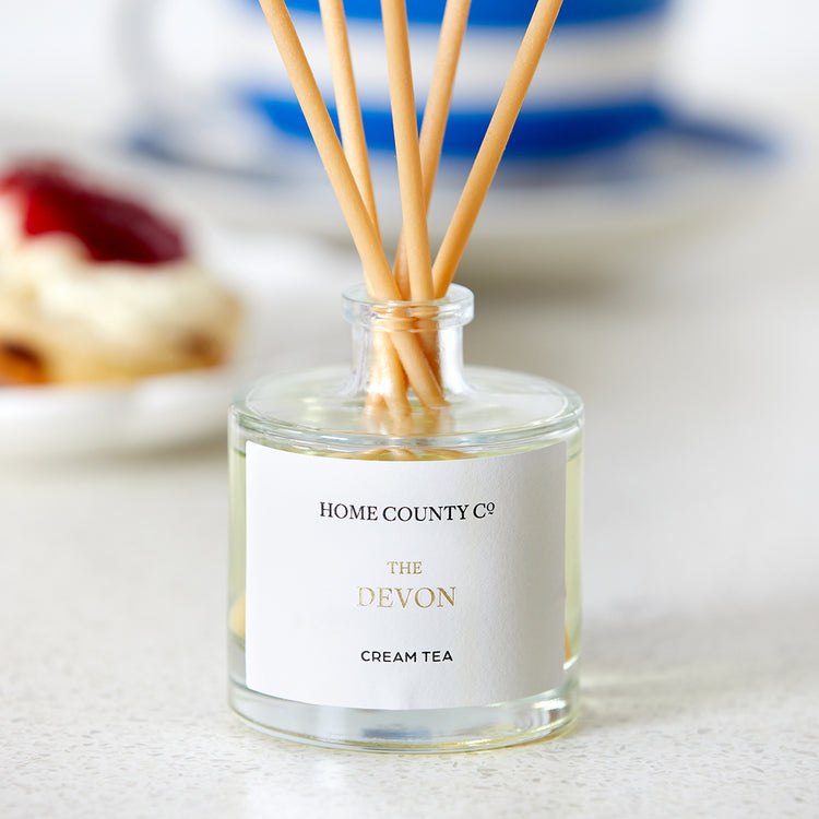 A Devon cream tea scented reed diffuser from the Home County Co. is shown in use on a kitchen worktop with a cream tea in the background
