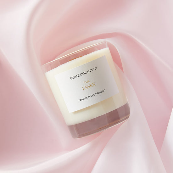 A Prosecco and Pomelo scented candle from the Home County Co is shown laid flat on a pink silk sheet