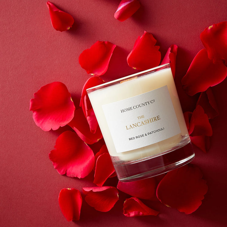 A red rose scented candle from the Home County Co is shown laid flat surrounded by red rose petals