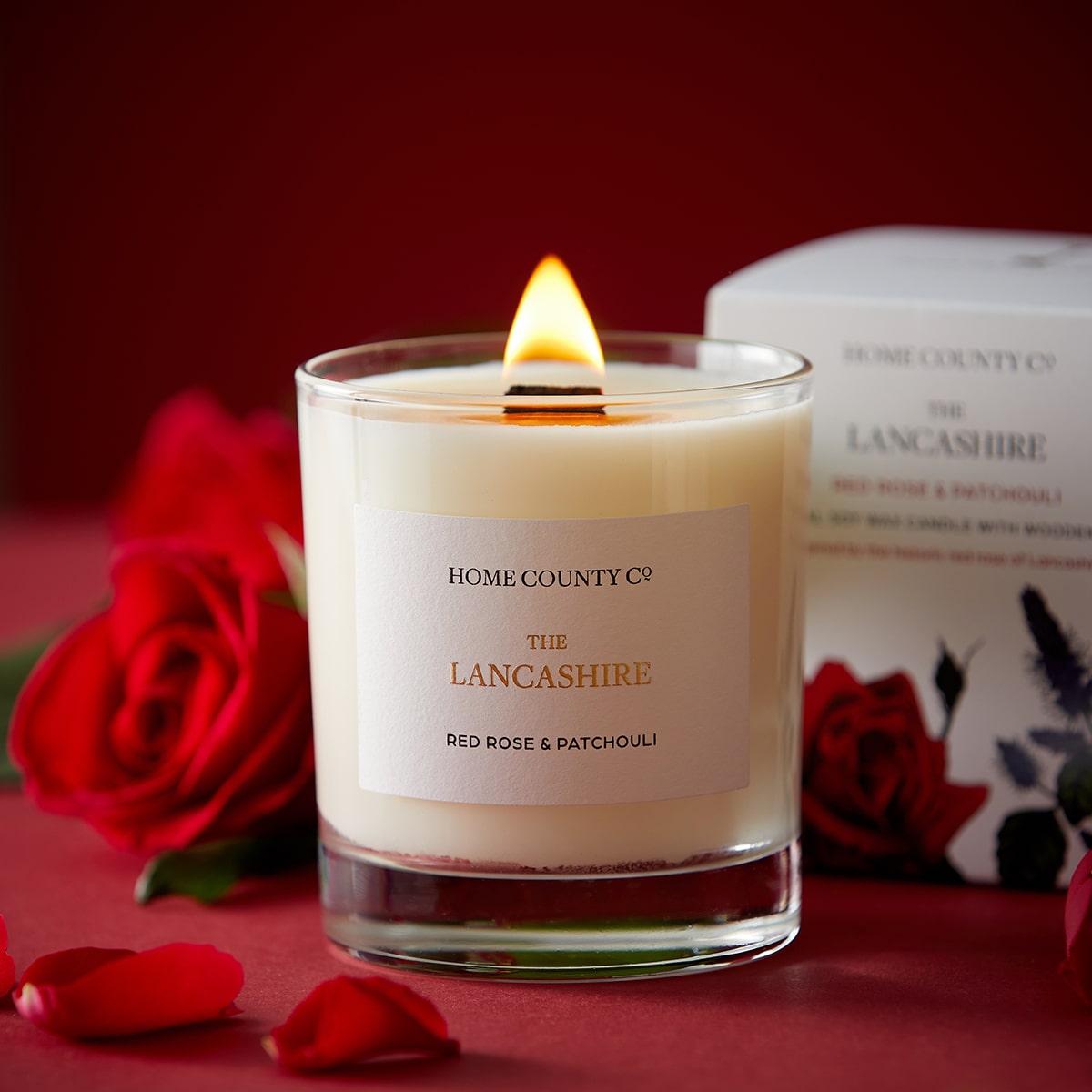 A red rose scented candle from the Home County Co is shown alight next to a red rose and its illustrated candle packaging