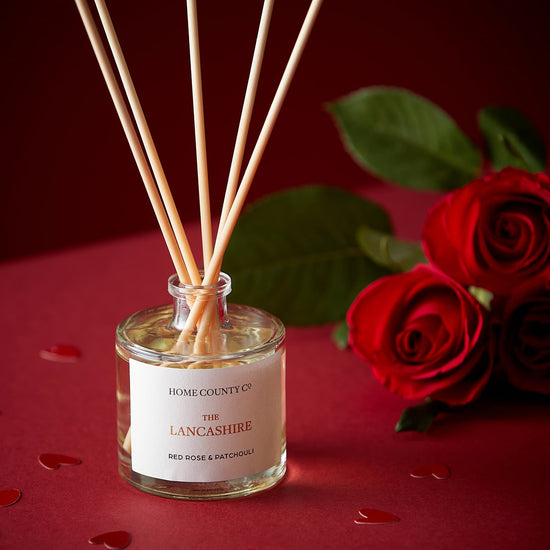 A Lancashire red rose and patchouli reed diffuser is shown next to fresh red roses